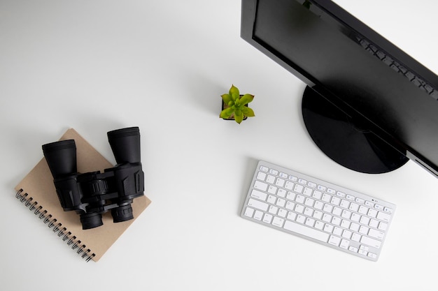 Binoculars lying on a notebook near a computer on a white table Top view flat lay