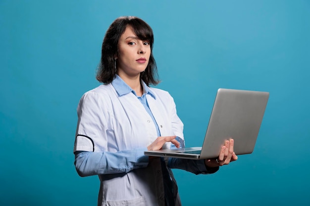 Biochemistry scientist standing on blue background having modern computer laptop while looking at camera. Microbiology specialist having portable electronic device looking at camera.