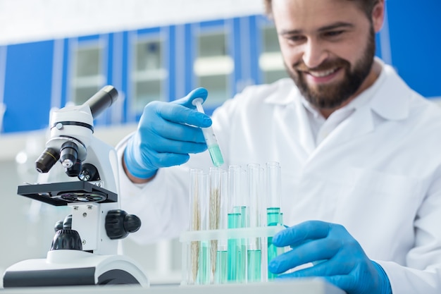 Biological project. Delighted nice smart biologist holding a syringe and putting blue liquid into test tubes while working on a scientific project