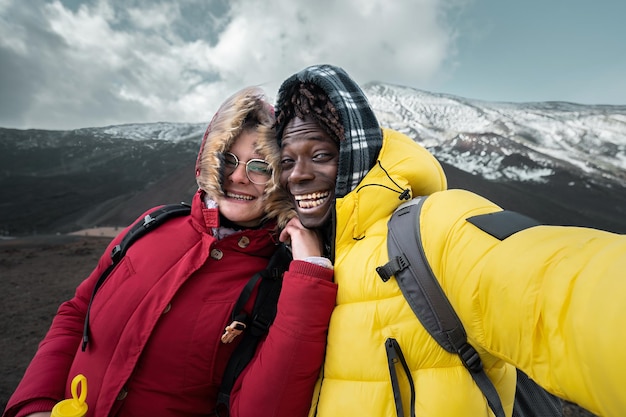 Biracial couple of young people hikers taking a smartphone selfie hugging on the peak of Vulcano Etna in Italy Europe tourism wanderlust and lifestyle concept