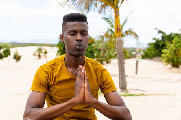 Biracial man with eyes closed practicing yoga meditation sitting on sunny beach, unaltered. Healthy lifestyle, wellbeing, relaxation, summer and vacation.