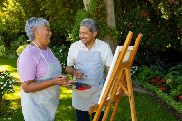 Biracial senior man looking at wife laughing while painting with watercolors on canvas in yard