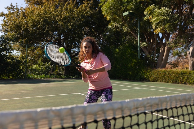 Biracial woman playing tennis returning ball over the net on sunny outdoor tennis court. Inclusivity, sport, healthy hobbies, competition, fitness and leisure time concept.