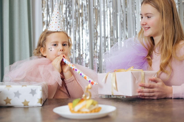 Birthday of 5 year old girl Two sisters are happy together surrounded by gifts birthday cake Birthday girl blows festive horn Holidays and birthdays Happy childhood