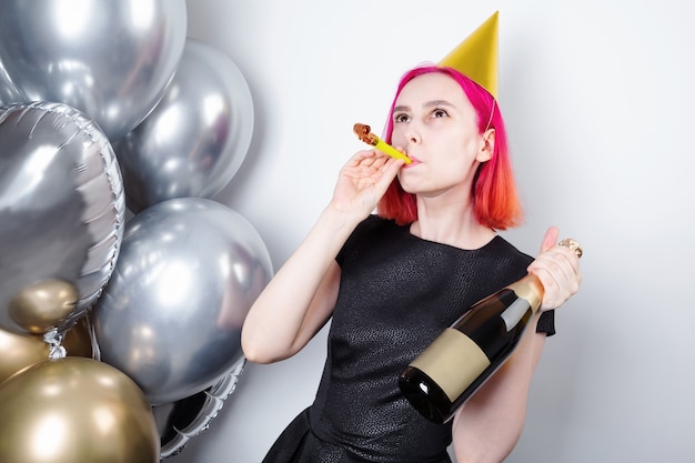 Birthday celebration. Young happy woman with pink hair with holiday cap and pipe holds bottle of champagne in hand, silver and gold balls