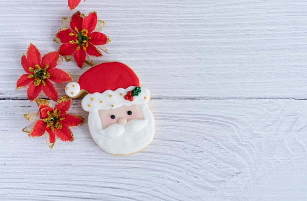 Biscuit with Santa's face on a white wooden background with red flowers decoration Top view Copy space