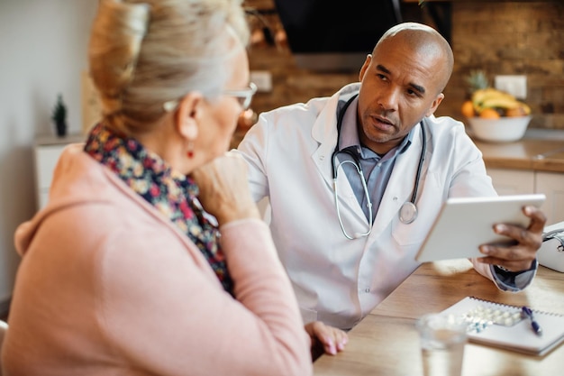 Blac doctor using digital tablet and communicating with mature woman during home visit