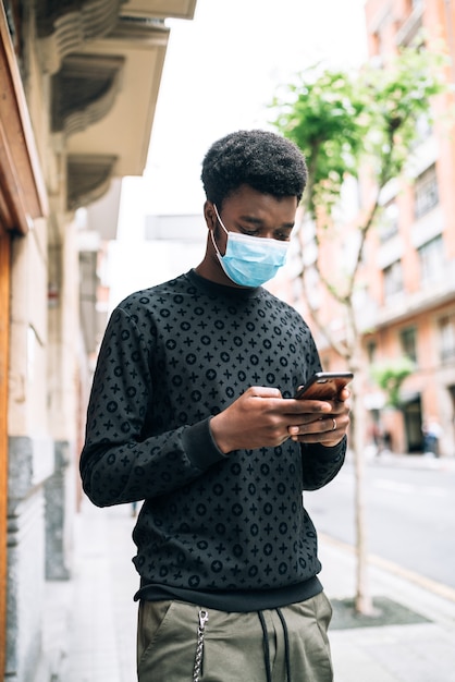 Black african american boy walking down the street with a blue face mask looking at his cell phone protecting himself from the covid-19 coronavirus pandemic