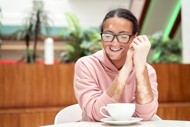 Black african american woman with vitiligo pigmentation skin problem indoor dressed pink hoodie glasses sitiing table indoor drink tea smiling Happy positive person waiting in restaurant