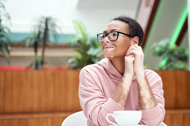 Black african american woman with vitiligo pigmentation skin problem indoor dressed pink hoodie glasses sitiing table indoor drink tea