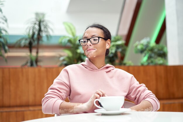 Black african american woman with vitiligo pigmentation skin problem indoor dressed pink hoodie glasses sitiing table indoor drink tea