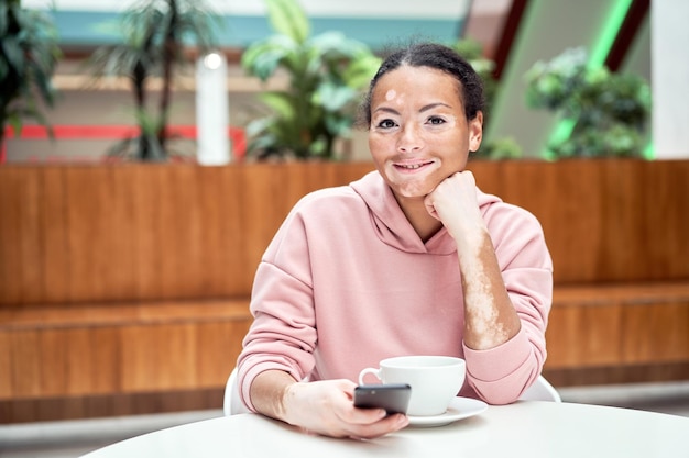 Black african american woman with vitiligo pigmentation skin problem indoor dressed pink hoodie sitting table using smartphone