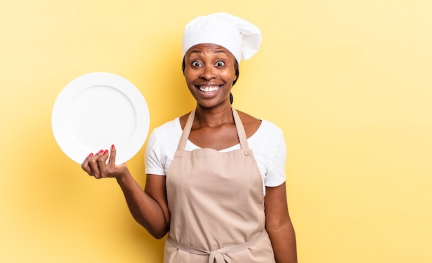 Black afro chef woman looking happy and pleasantly surprised, excited with a fascinated and shocked expression. empty plate concept