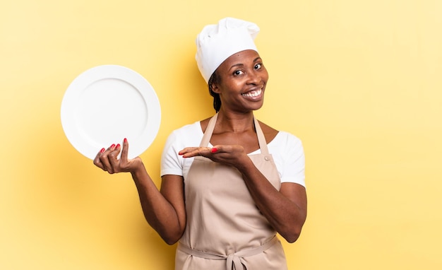 Black afro chef woman smiling cheerfully, feeling happy and showing a concept in copy space with palm of hand. empty plate concept