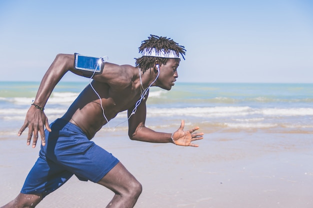 Black athlete running man - male runner at the beach listening to music on smartphone. Jogger training with smart phone armband,