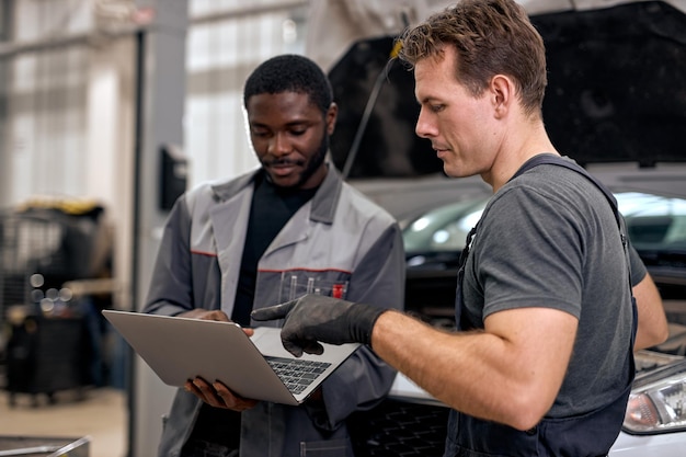 Black auto mechanic and caucasian repairman checking diagnostics results on laptop computer explains...