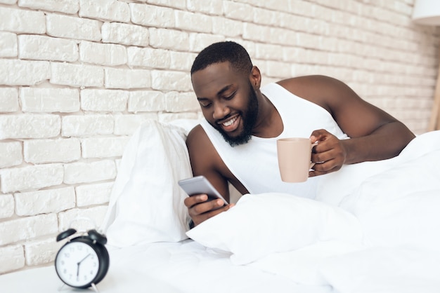 Black, awakened man drinks coffee in bed browsing web.