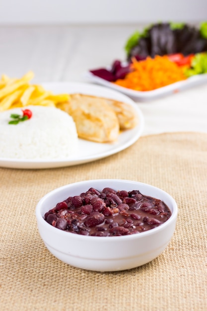 Black beans on white plate on raw cotton dish towel with a full plate with rice chicken and fries behind