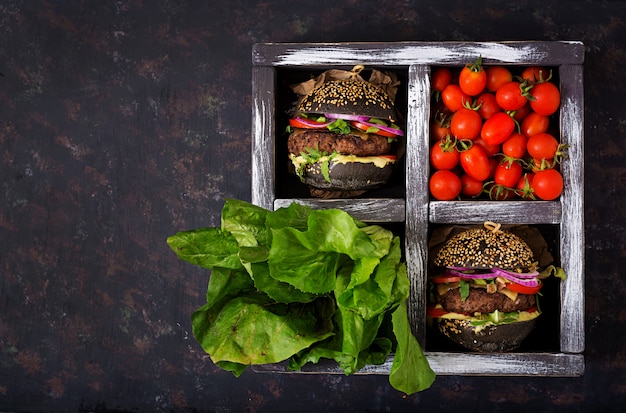 Black big sandwich -  black hamburger with juicy beef burger, cheese, tomato,  and red onion in box on black surface.  Flat lay. Top view