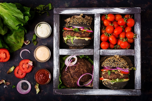 Black big sandwich -  black hamburger with juicy beef burger, cheese, tomato,  and red onion in box on black surface.  Flat lay. Top view