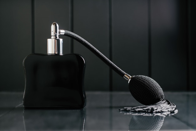 Black bottle of eau de toilette or perfume with long tassel spray pomp stands on dark background on reflective surface. Copy space