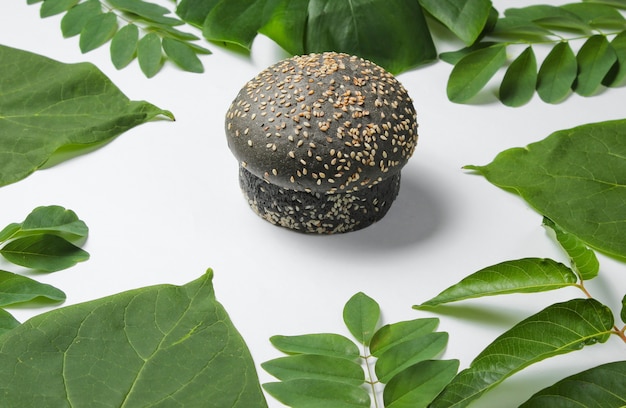 Black bun with sesame seeds on a white background with green tropical leaves