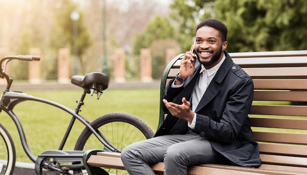 Black business professional is using his phone in a park seated on a bench with a bicycle beside him