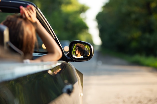 Black cabriolet on the road on a sunny day. In the side mirror is reflection of the dark-haired young man with beard. .