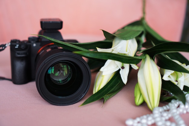 Black camera with a lens and white lilies and a necklace of pearls on a pink background. Selective focus. No visible logos and brands