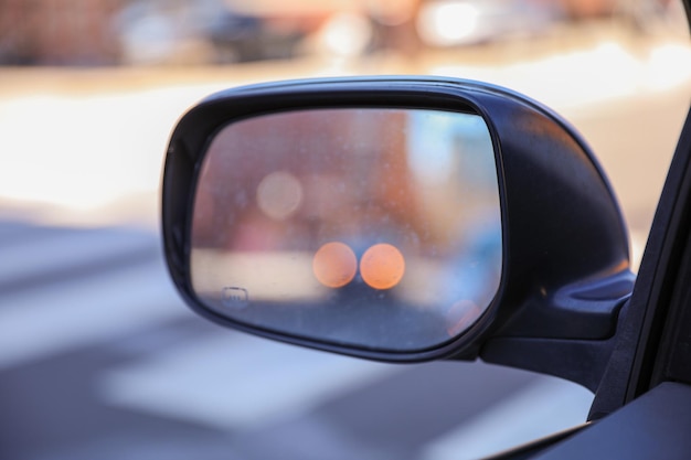 A black car's side mirror has orange lights in the background.