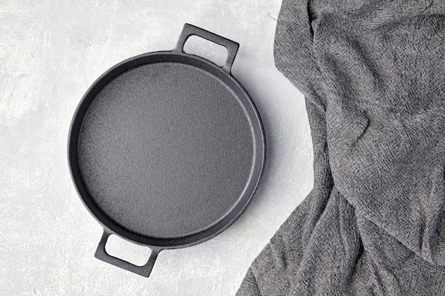 Black cast iron empty frying pan and gray kitchen towel on a gray concrete background