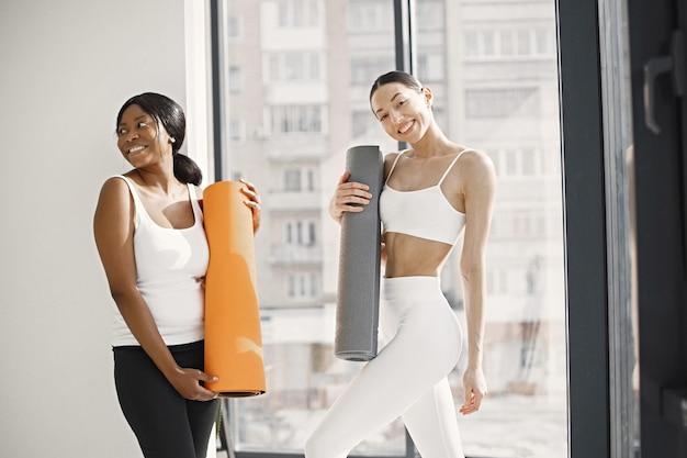 Black and caucasian women standing with rolling mats in studio with big windows