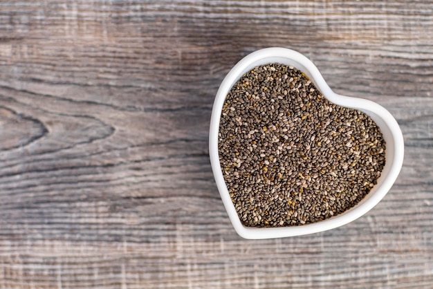 Black chia seeds in a white heart shaped bowl on wooden background Superfood for healthy eating and diet