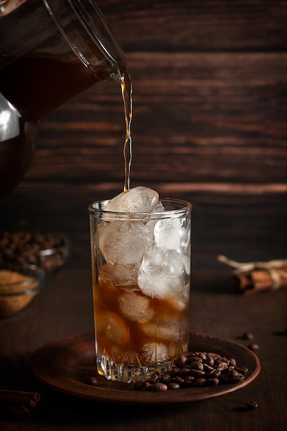 Black coffee being poured from jar into glass with ice cubes on plate with roasted beans on table