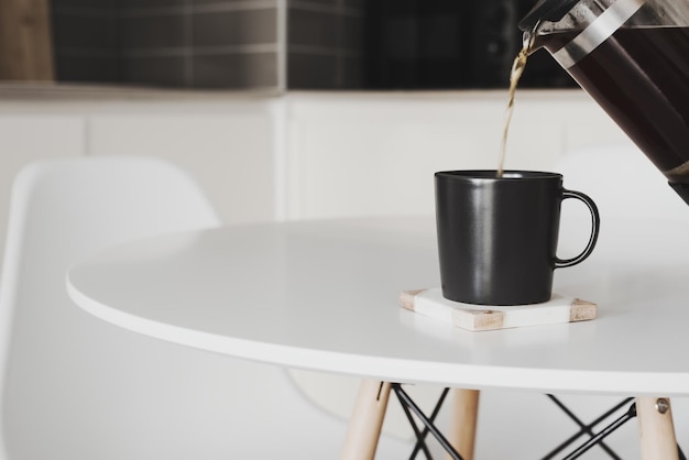 Black coffee brewed in a french press coffee maker pouring into a black mug. Homemade coffee