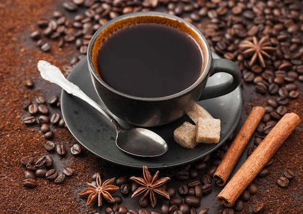 Black coffee in ceramic cup with cinnamon and cane sugar with anise star and silver spoon on ground coffee and beans background