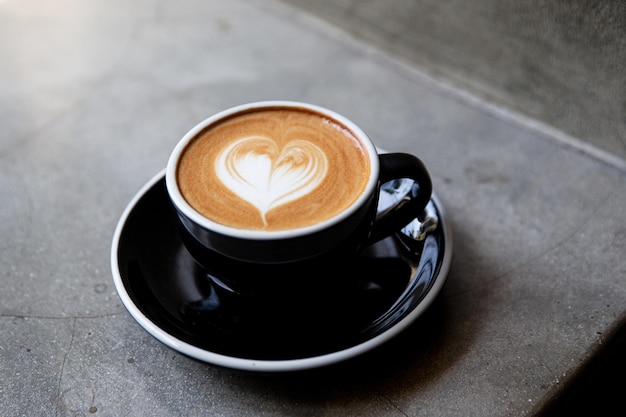 Black cup of cappuccino with latte art of heart shape on saucer on concrete background