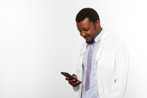 Black doctor man with small beard in white coat looks at smartphone isolated on white background