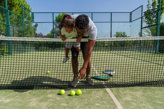 Black father and daughter taking ball from ground on tennis court