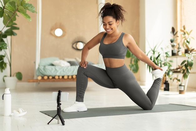 Black female fitness blogger stretching in front of smartphone at home shooting video for her