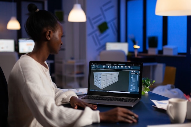 Black freelancer architect working in 3D software to elaborate container design sitting at desk in business office at midnight