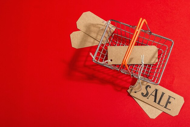 Black Friday. Sale tags with a shopping basket on the red background. Zero waste shopping concept, trendy hard light, dark shadow, flat lay, top view