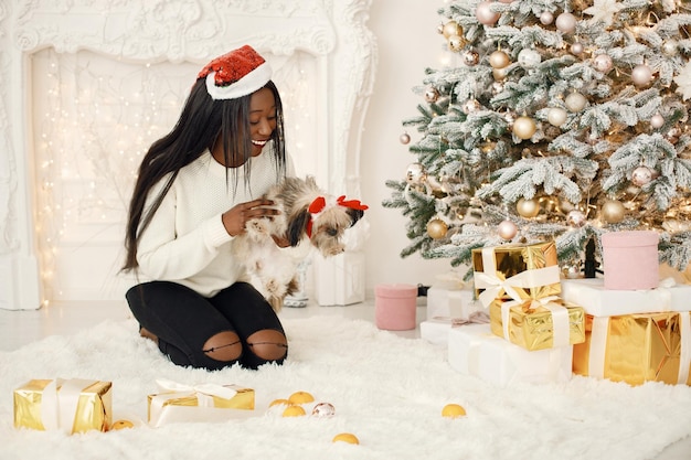 Black girl in Santa's hat sitting near Christmas tree holding a little dog