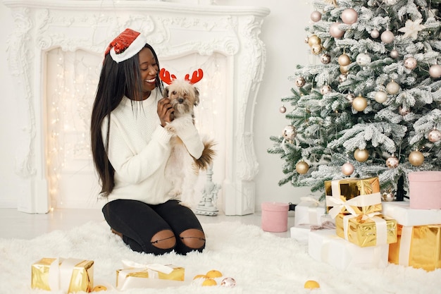 Black girl in Santa's hat sitting near Christmas tree holding a little dog