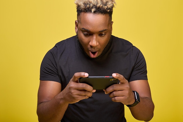 Black guy with his mouth open looks at mobile phone screen on yellow background