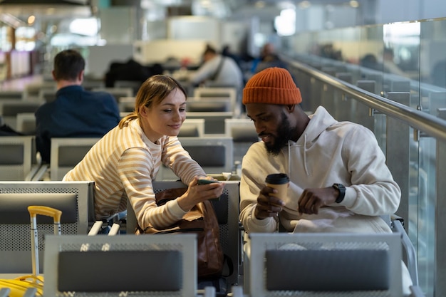 Black hipster male and caucasian pretty female looking at phone while waiting for flight at airport