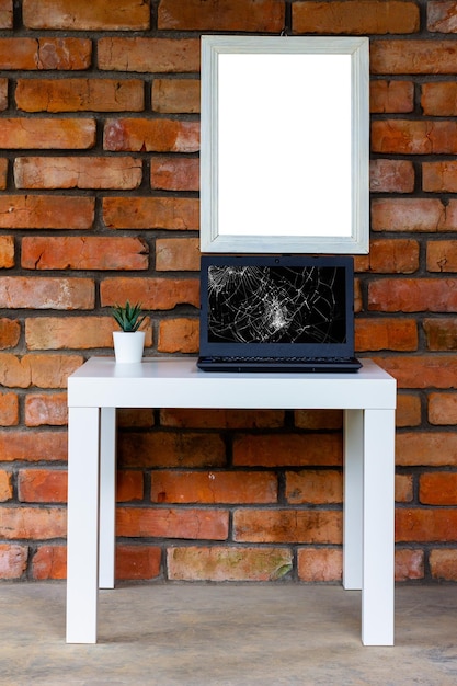 A black laptop with a broken screen on a white table against an old brick wall with a white mockup in a white cardboard frame