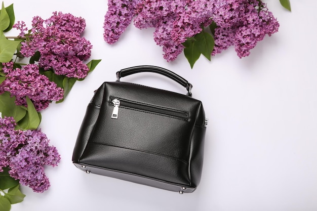 Black leather bag with branches of blooming lilacs on white background Spring fashion concept Top view Flat lay