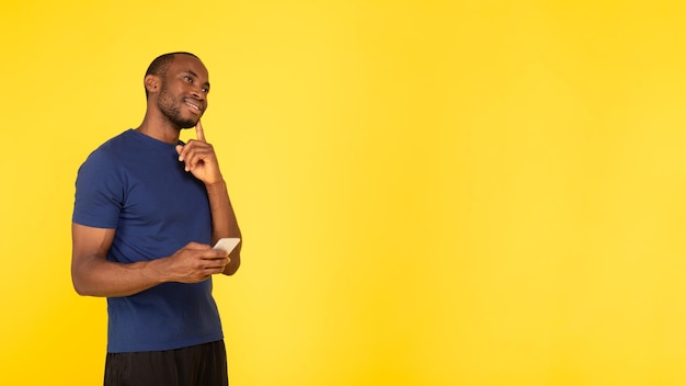 Black Male Thinking Holding Smartphone Looking Aside Over Yellow Background