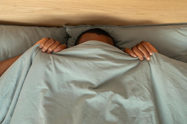 Black Man Hiding Covering Face With Blanket Lying In Bedroom